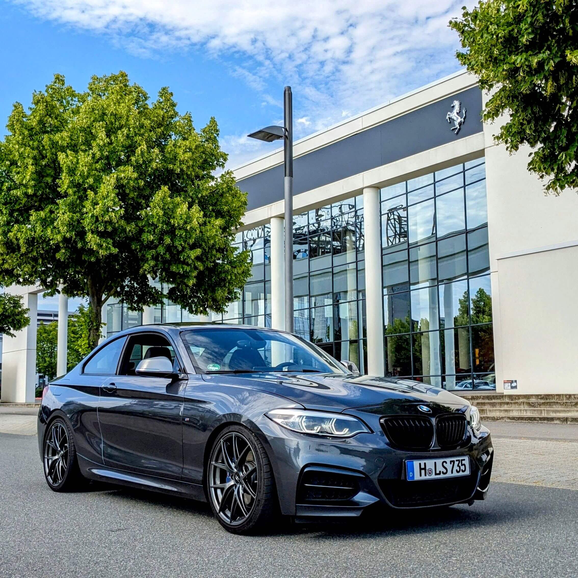Ein dunkelgraues BMW Coupé steht auf der Straße vor einem modernen Glasgebäude mit Ferrari-Logo