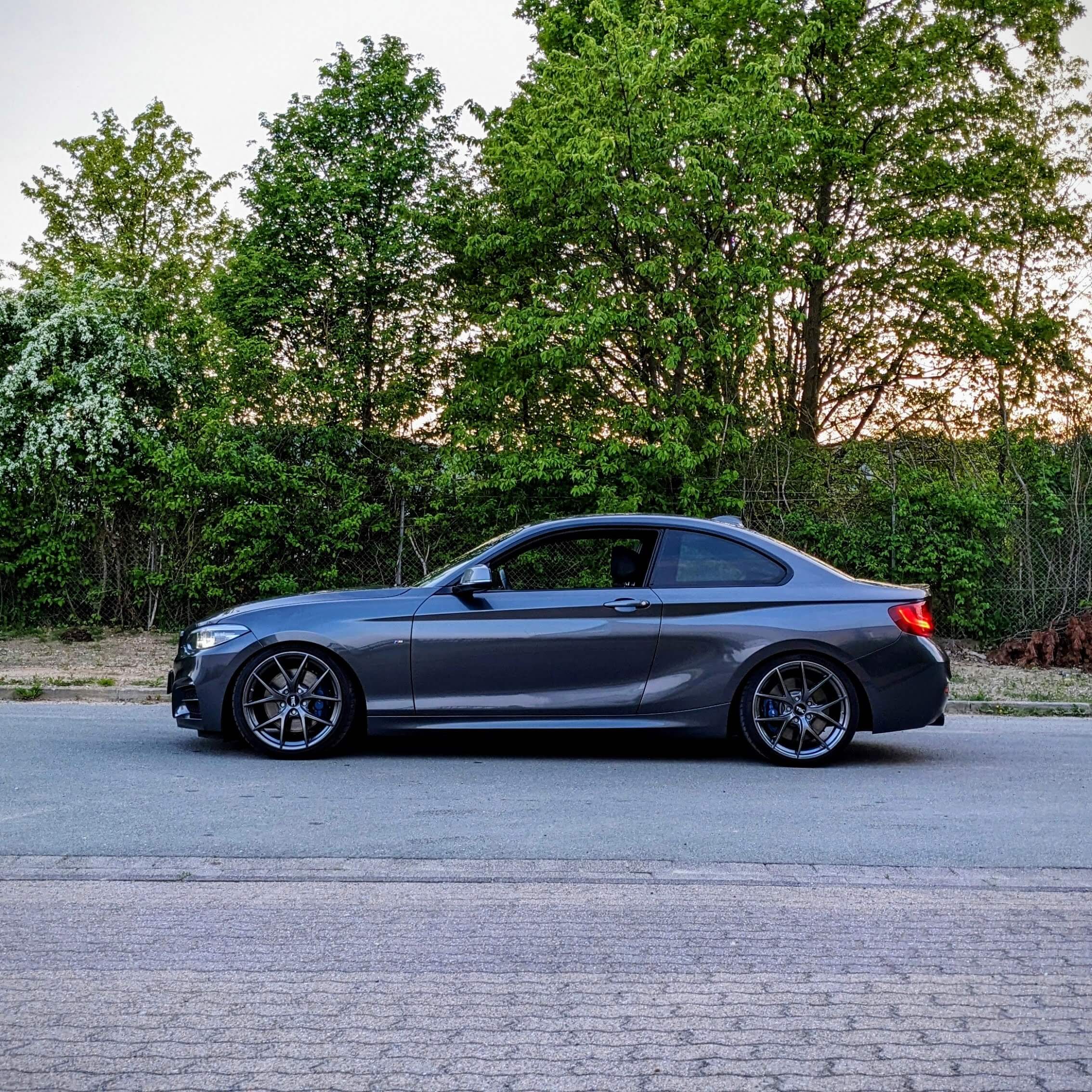 Ein dunkelgraues Coupé steht auf einer Straße, dahinter sind grüne Bäume und Büsche zu sehen