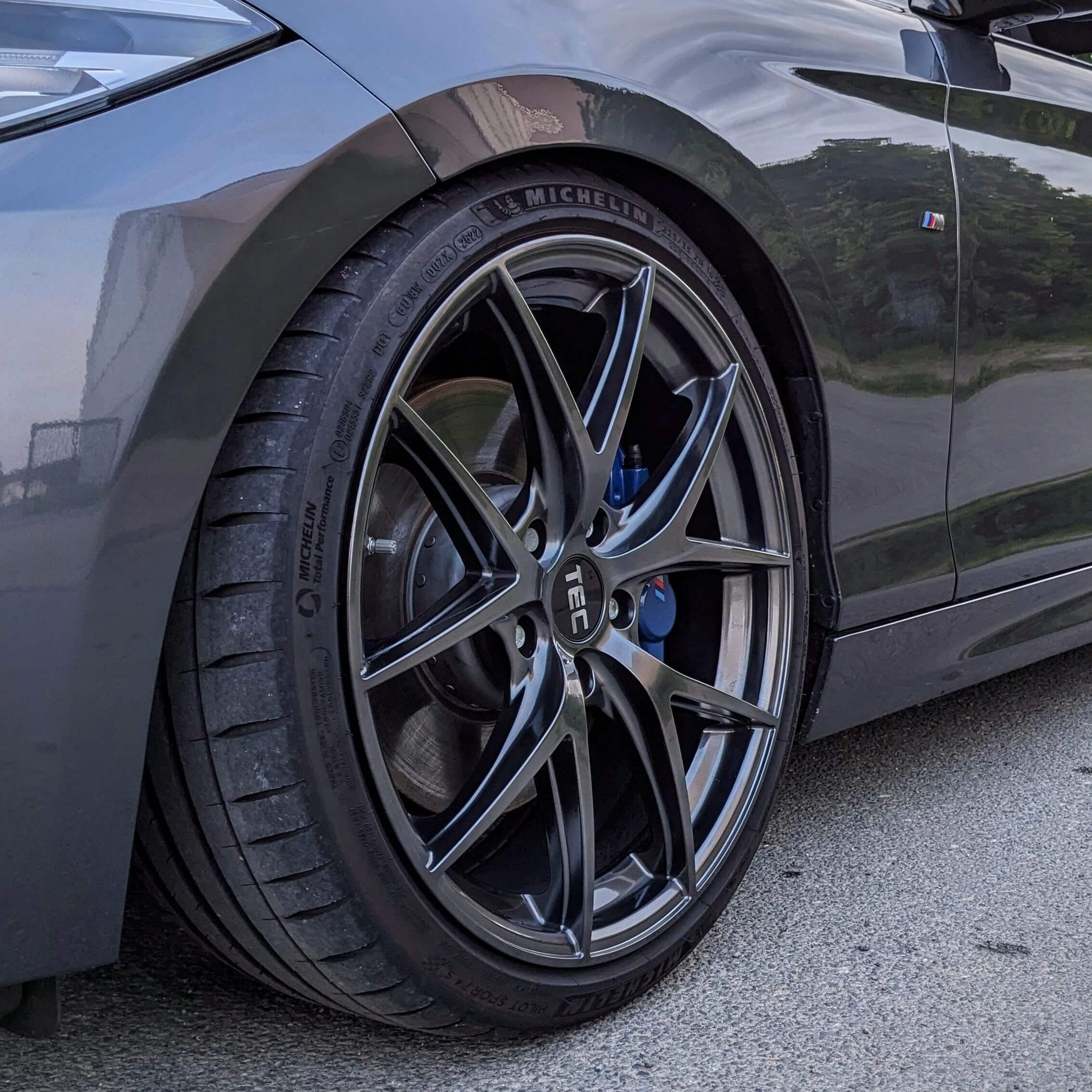 Nahaufnahme eines vorderen Autoreifens mit schwarzen Alufelgen, Michelin-Reifen und M-Emblem an einem glänzend grauen Kotflügel