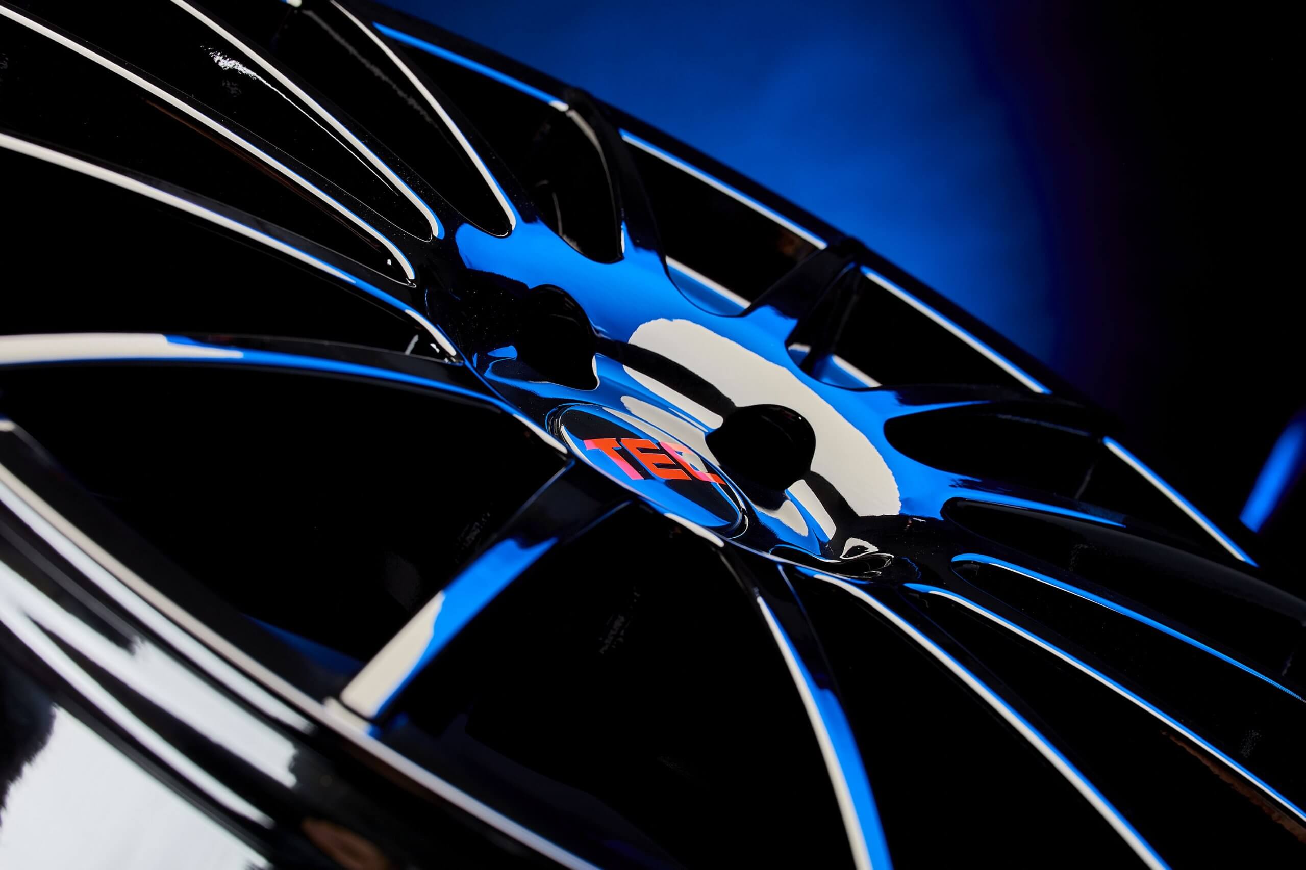 Eine glänzend schwarze TE37-Felge mit dünnen Speichen und rotem Logo vor blauem Hintergrund