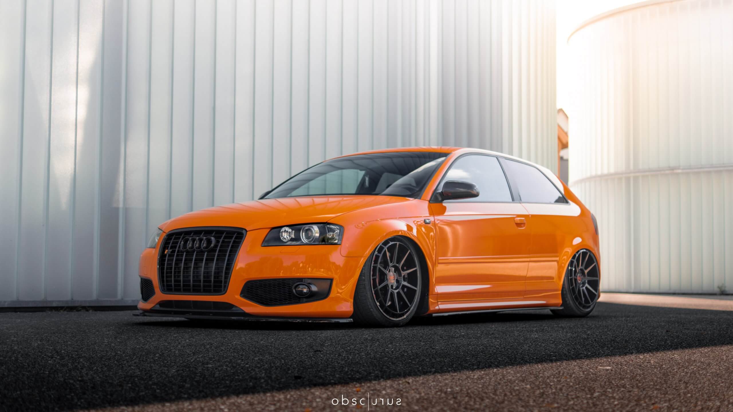 Ein leuchtend orangefarbener, getunter Audi Hatchback mit schwarzen Felgen steht vor einem modernen weißen Industriegebäude