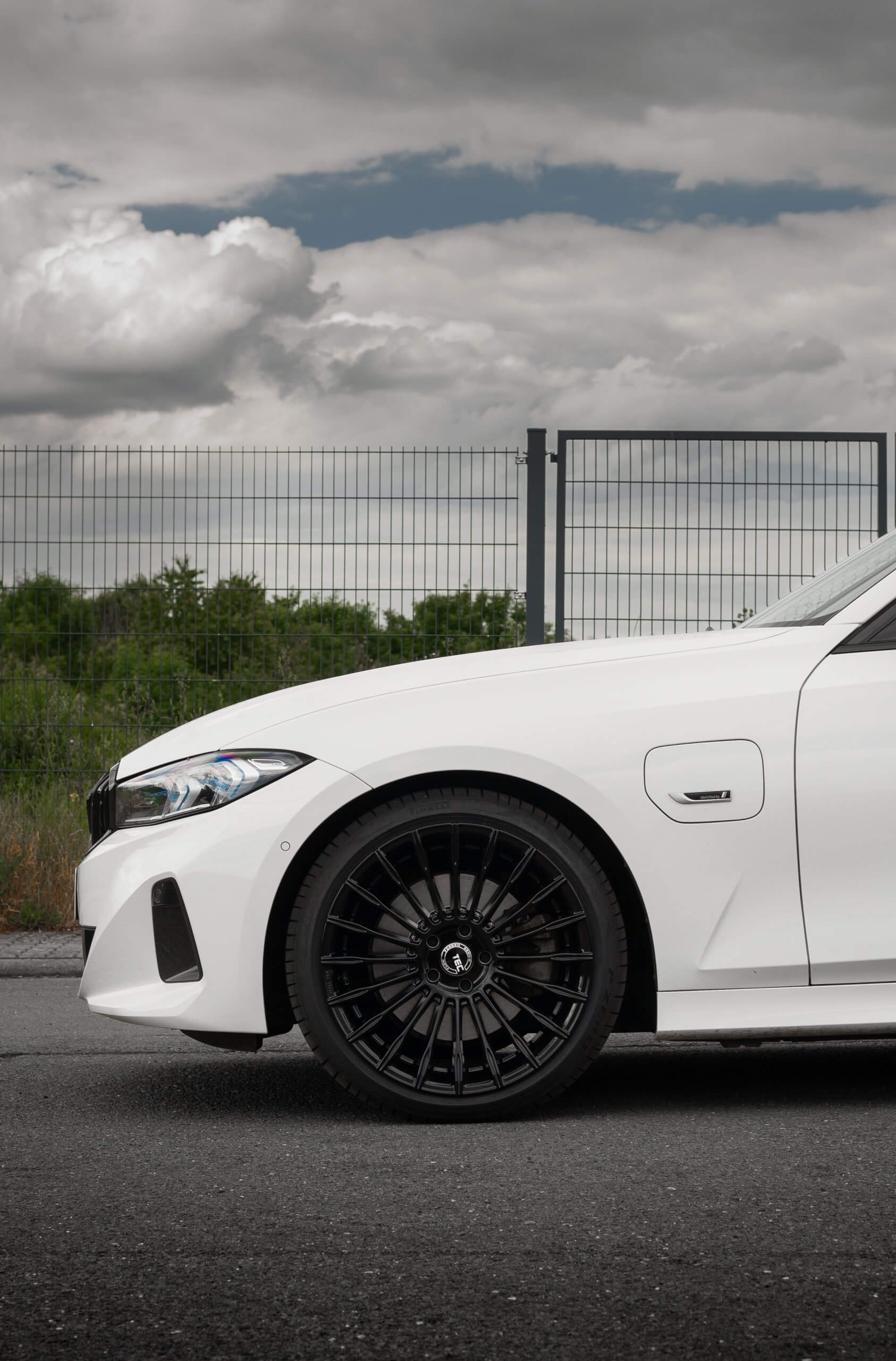 Ein weißes Auto von der Seite, vorn das Rad sichtbar, auf Asphalt bei einem Metallzaun und etwas Grün unter bewölktem Himmel