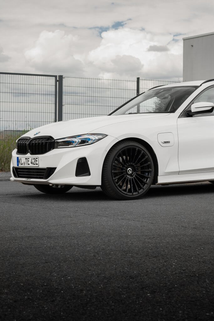Ein weißer BMW steht auf Asphalt nahe einem Zaun, zu sehen ist die Frontlinke Seite mit Rad und darüber ein bewölkter Himmel