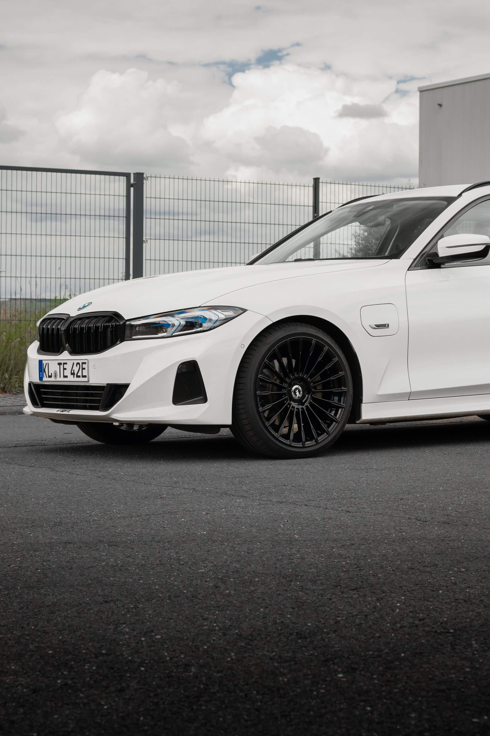 Ein weißer BMW steht auf Asphalt nahe einem Zaun, zu sehen ist die Frontlinke Seite mit Rad und darüber ein bewölkter Himmel