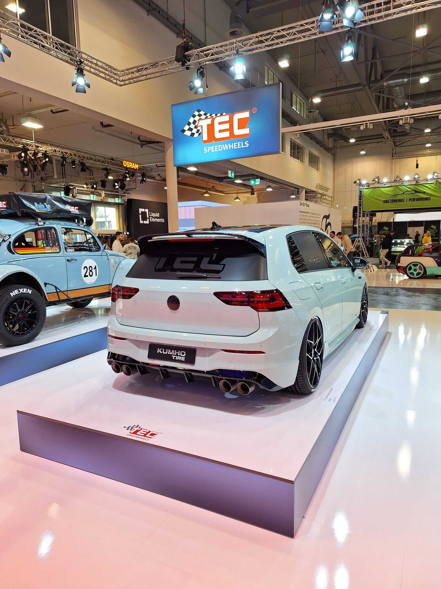 Ein weißer TEC Speedwheels steht rückseitig sichtbar auf einer Automesse, umgeben von weiteren Autos und Ausstellungsständen