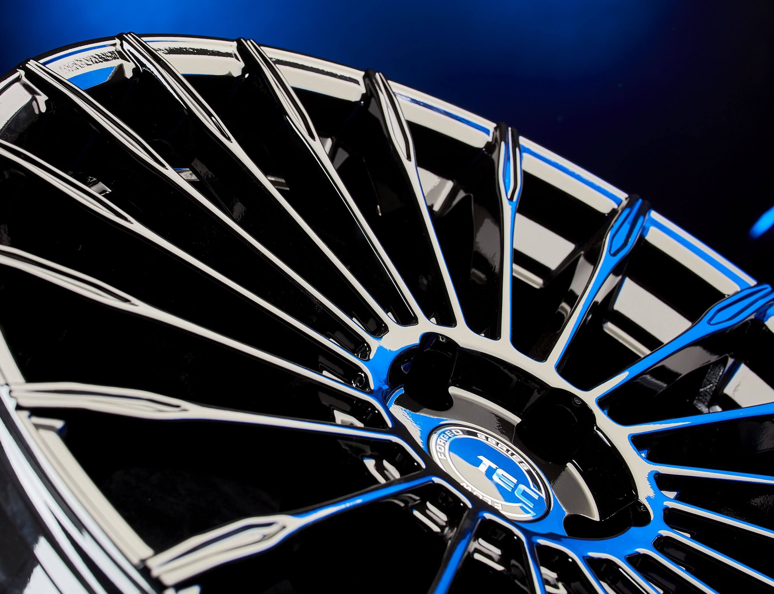 Eine schwarz-blaue Alufelge mit glänzendem, filigranen Speichendesign vor dunkelblauem Hintergrund
