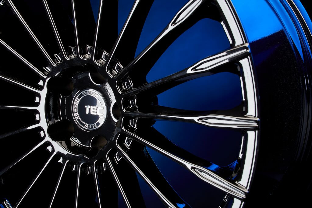 Nahaufnahme einer glänzenden schwarzen TEC-Alufelge vor blauem Hintergrund mit detaillierten Speichen und zentralem Logo