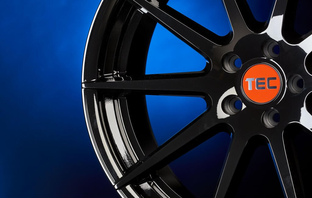 Nahaufnahme einer glänzend schwarzen Alufelge mit orangefarbenem TEC-Schriftzug in der Mitte vor blauem Hintergrund