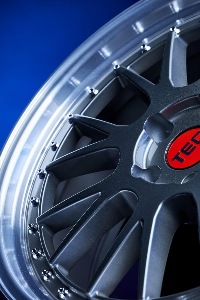 Nahaufnahme einer metallischen Autofelge mit dunklen Speichen, silbernen Bolzen und roter Nabenkappe vor blauem Hintergrund