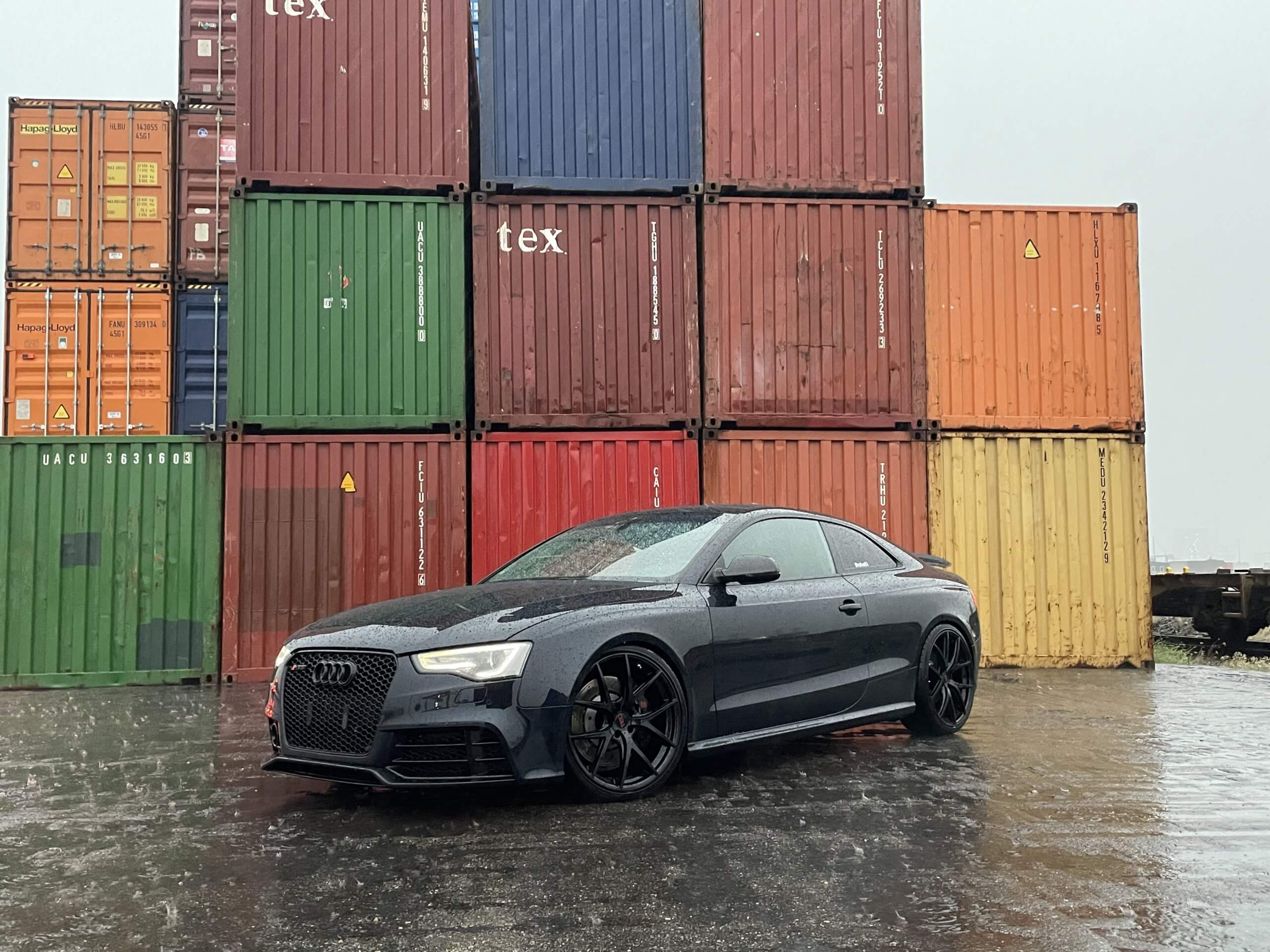 Ein schwarzes Audi Coupé steht auf nassem Asphalt vor gestapelten bunten Schiffscontainern an einem wolkigen Tag