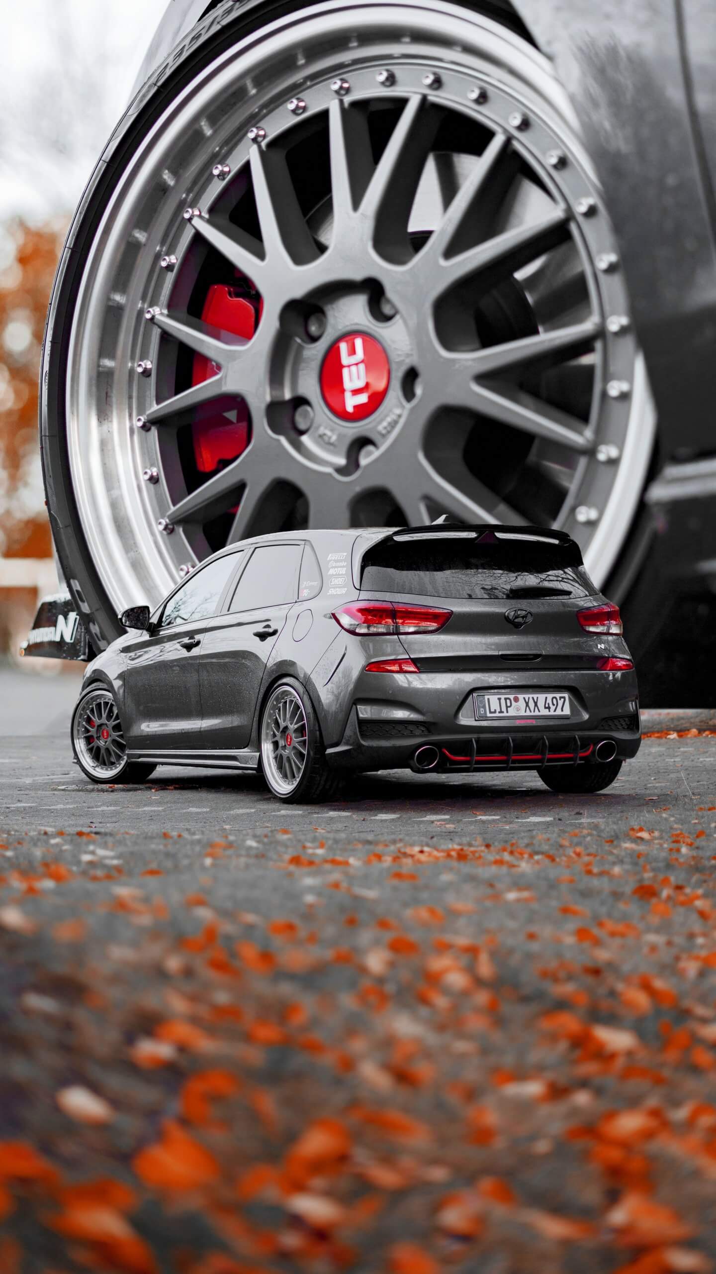 Ein schwarzes Auto steht auf Herbstlaub, im Hintergrund ist ein riesiges individuelles Rad mit auffälliger Felge zu sehen
