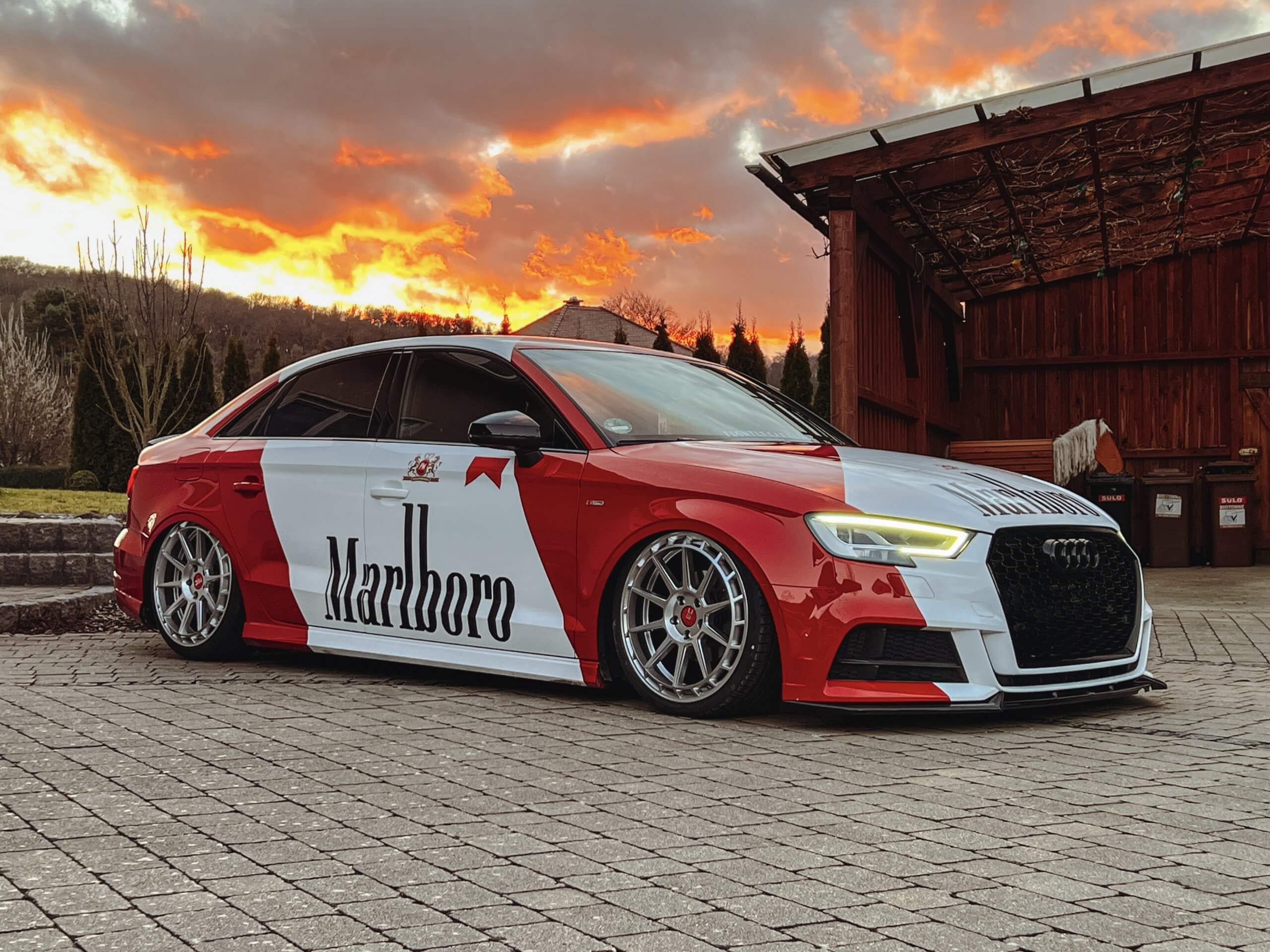 Ein rot-weißer Audi im Marlboro-Look steht bei Sonnenuntergang auf einer Auffahrt aus Ziegelsteinen unter dramatischen Wolken