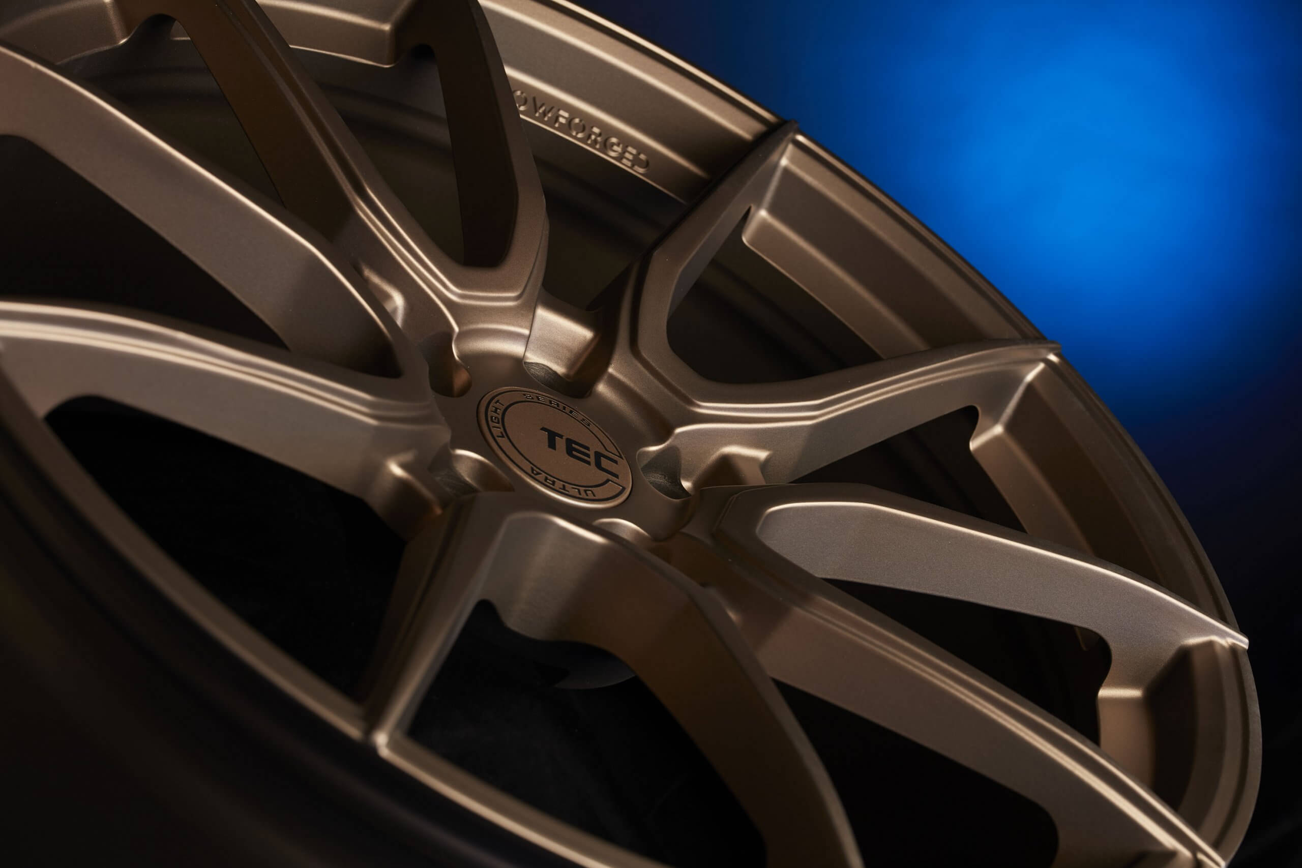 Nahaufnahme einer bronzefarbenen flowforged TEC-Felge mit Logo vor blauem Farbverlauf-Hintergrund