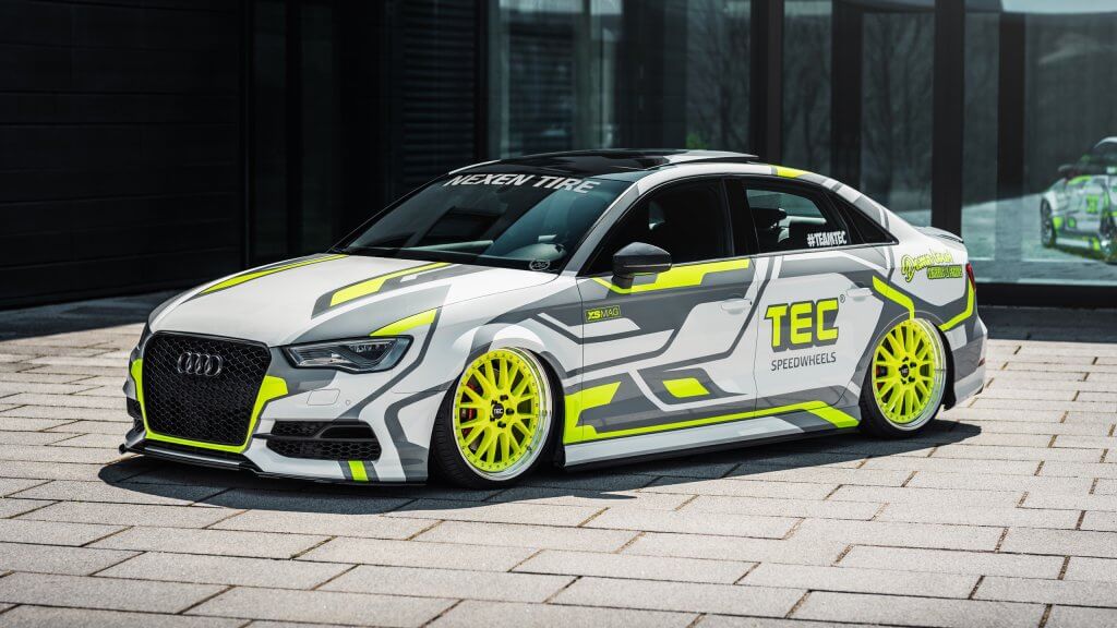 Ein weißer Audi mit neongelben Felgen und grafischen Aufklebern steht vor einem modernen Gebäude mit Glasfassade