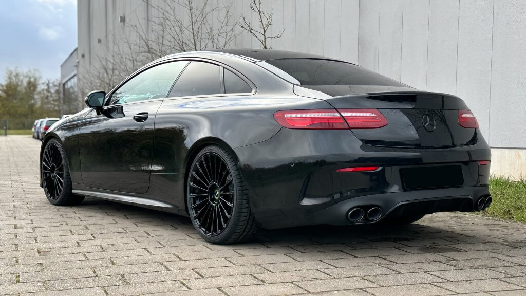 Ein elegantes schwarzes Coupé steht auf einem gepflasterten Platz neben einem weißen Gebäude, von hinten seitlich betrachtet