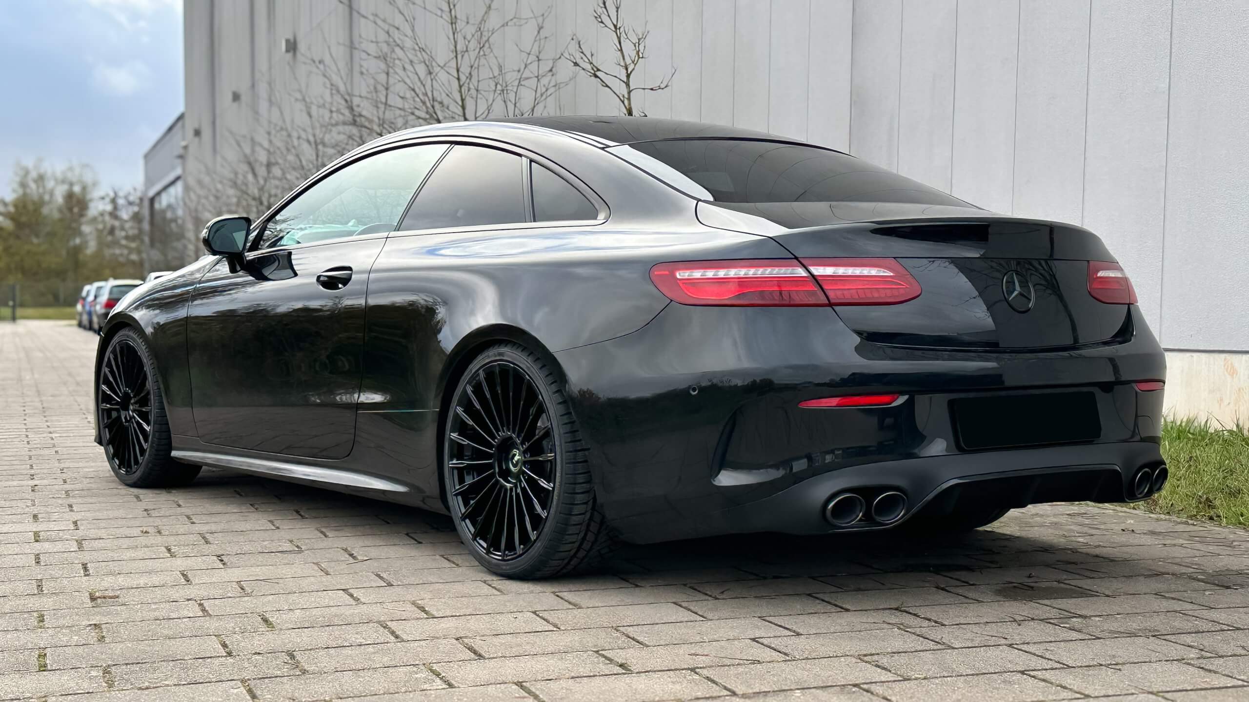 Ein elegantes schwarzes Coupé steht auf einem gepflasterten Platz neben einem weißen Gebäude, von hinten seitlich betrachtet