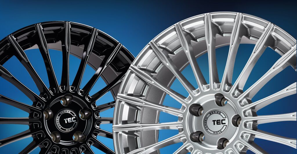 Zwei Alufelgen mit Speichendesign und TEC-Logo, eine glänzend schwarz, eine silberfarben, liegen nebeneinander auf blauem Hintergrund